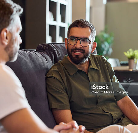 Two men sat on a sofa smiling at each other