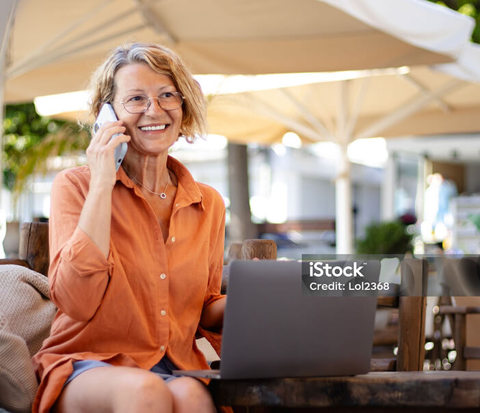 Photo of a woman in an outdoor dining area, in front of her laptop, speaking on the phone