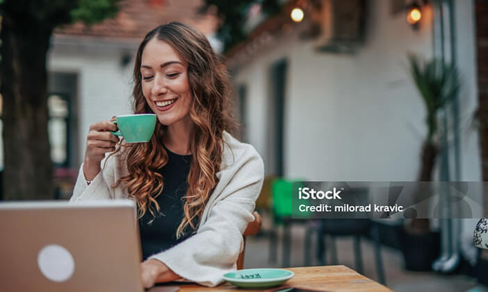 Photo of a young woman at a cafe, sipping her coffee while smiling at her laptop
