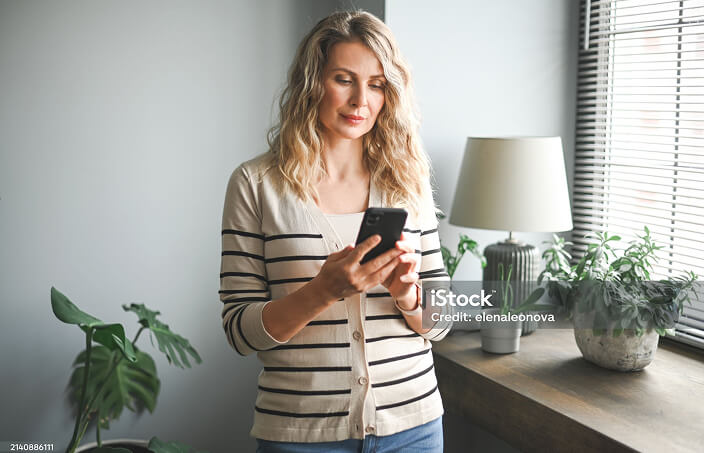 Photo of a woman at home, standing by a window, using her mobile phone